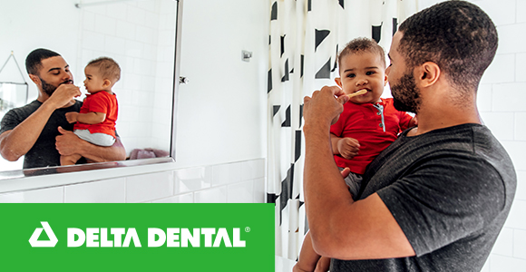 A dad brushing his little boy's teeth in front of a mirror.